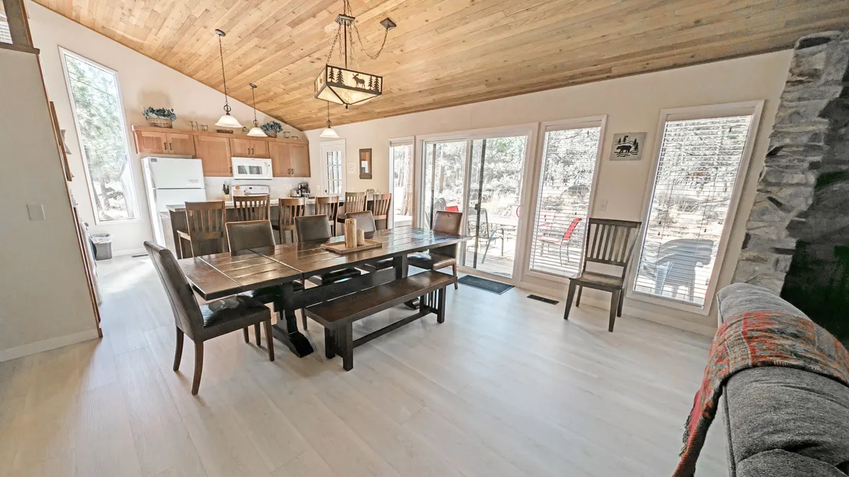 A harmonious interior featuring a dining table, large windows filling the room with natural light, a visible kitchen area, and a stone fireplace. The layout suggests seamless flow between dining, cooking, and lounging—perfect for multi-generational families or friends to connect.