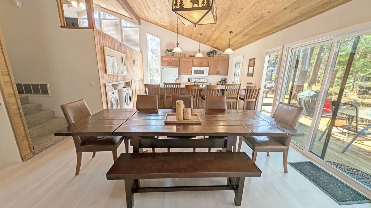 This image features a spacious open-plan area that showcases a large wooden dining table at the center, with the open kitchen area visible behind it. The space is bathed in natural light from generous windows, and a rustic light fixture adds a warm, inviting touch. The scene hints at the ease of moving between kitchen, dining, and living zones, making it ideal for gatherings.