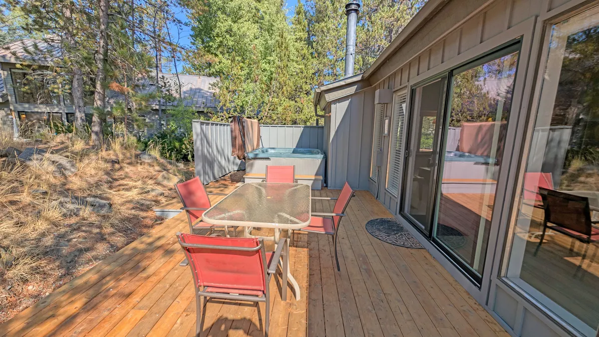 This exterior photo captures a spacious wooden deck surrounded by tall trees. The deck features an outdoor dining table and a prominent hot tub, with ample seating for relaxing. The natural setting creates a secluded, tranquil atmosphere perfect for outdoor enjoyment at any time of day.