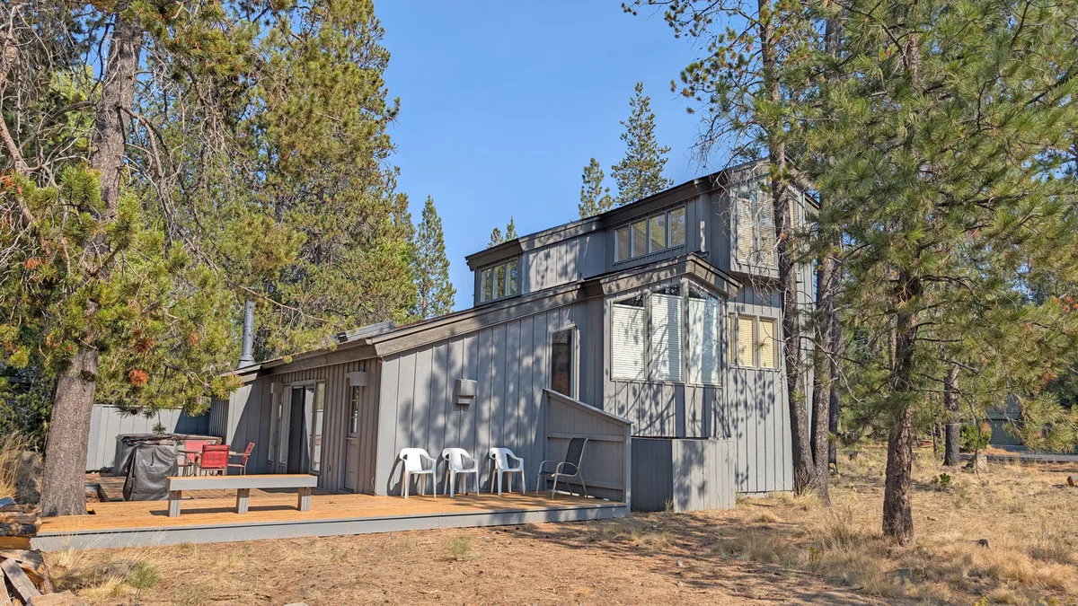 An inviting modern cabin exterior set amid tall pine trees, with a spacious deck featuring comfortable seating. The surrounding greenery and tranquil atmosphere offer a sense of adventure and relaxation, perfect for gathering after a day outdoors.