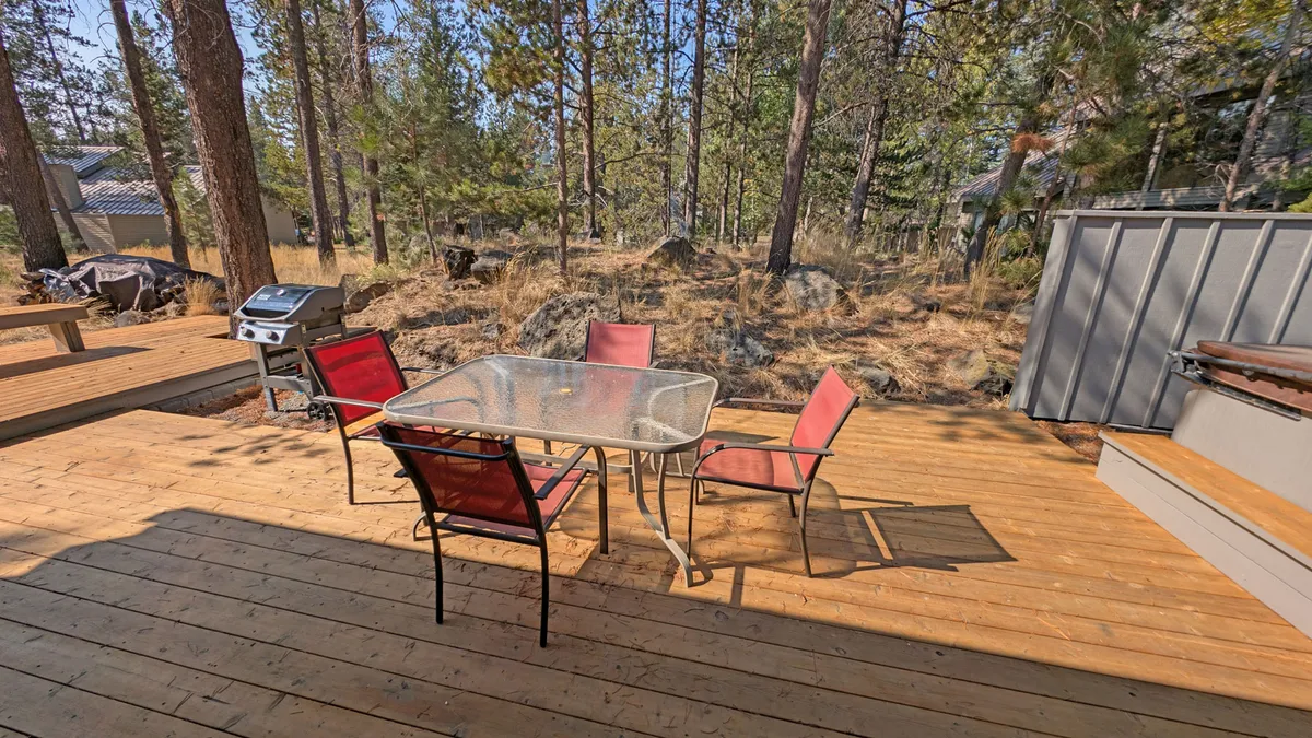 A cozy wooden deck equipped with a glass-top table, red chairs, a grill, and surrounded by lush trees. The setting is ideal for outdoor dining, lounging, and soaking in the peaceful natural ambiance.