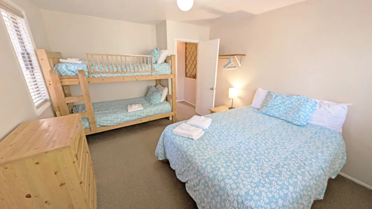 A bright, family-friendly bedroom with a double bed and a bunk bed, complemented by a nightstand with a lamp and a dresser. The furnishings and layout offer both comfort and flexibility, accommodating families with children or a group seeking versatile sleeping arrangements.