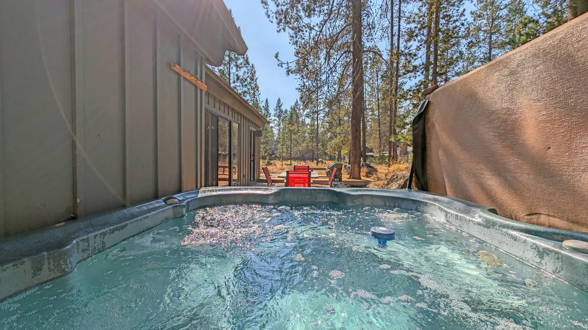 The exterior of a spacious, multi-level home nestled among tall Oregon pines, with a private hot tub surrounded by red Adirondack chairs on a sun-dappled deck. The setting is peaceful and inviting, perfect for relaxing and connecting with nature.