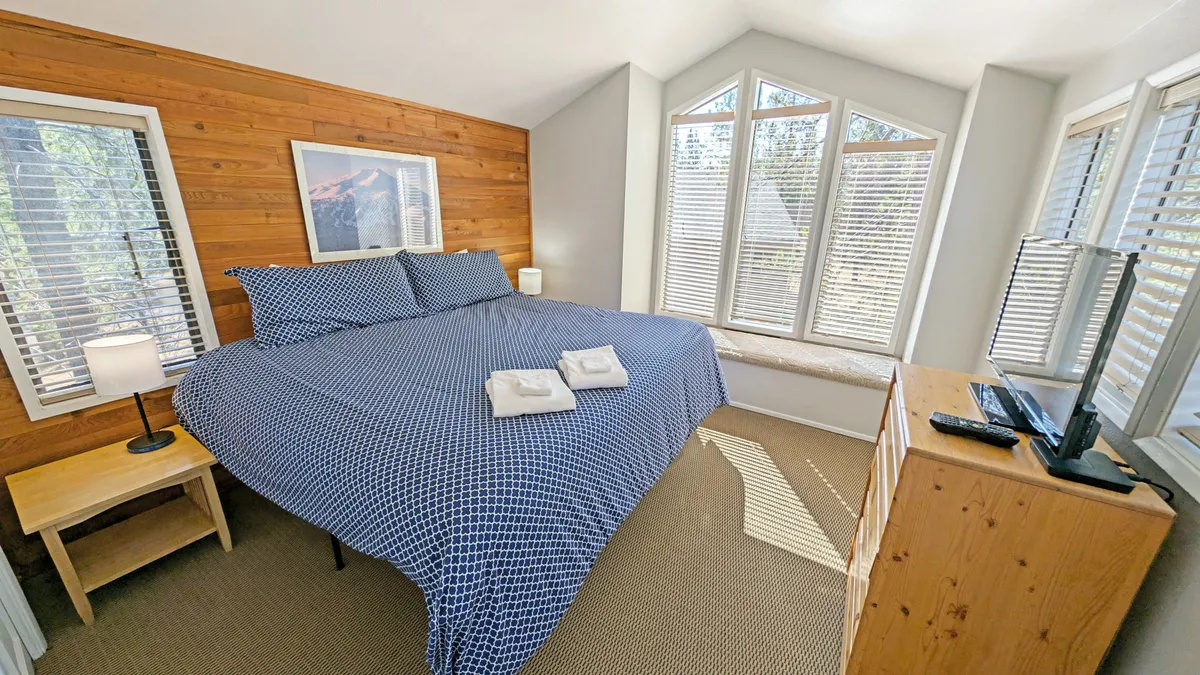 This image depicts a tranquil king bedroom featuring a bed with blue patterned linens, large windows inviting in natural light, a wooden accent wall, and a nightstand with a lamp. The room exudes a calm, serene atmosphere, perfect for relaxation and privacy.
