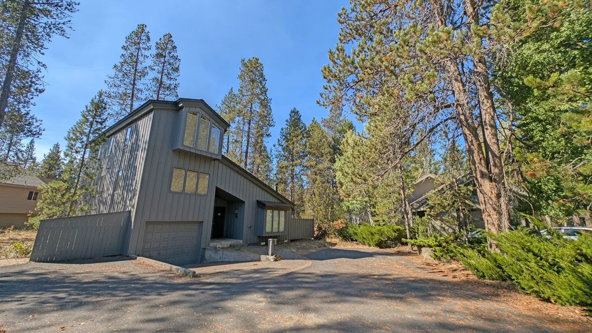This exterior image highlights the modern architecture of the home, surrounded by tall evergreen trees and set against a clear sky. The front porch and tranquil forest setting evoke a strong sense of peaceful seclusion.
