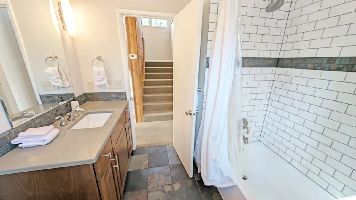 A bright, functional shared bathroom with a vanity and sink, spacious shower area, and a staircase leading upwards. Natural light fills the space, creating a fresh and inviting mood suitable for both kids and adults.