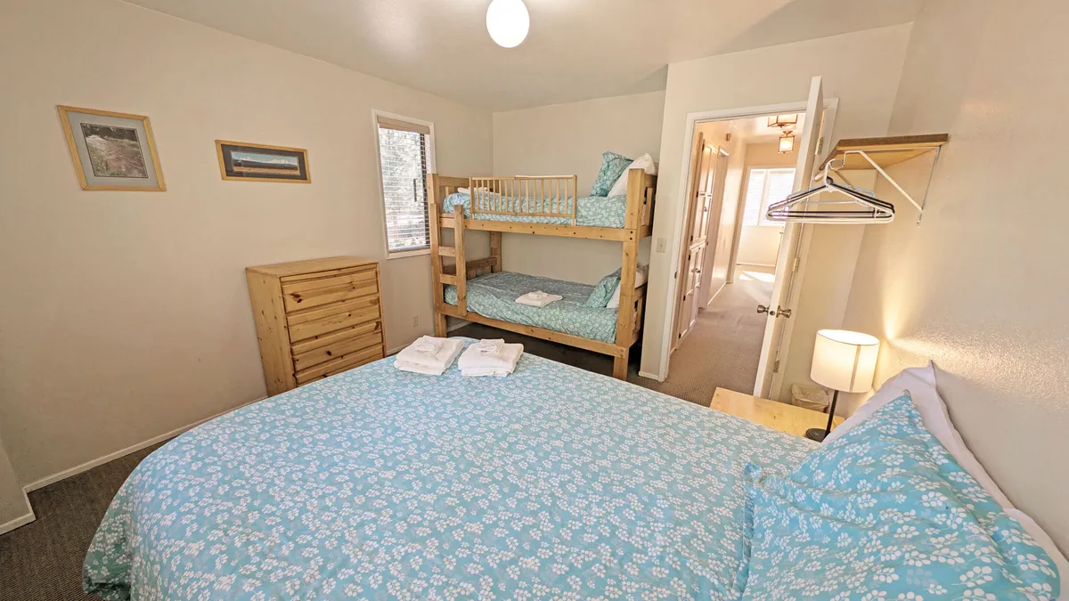 A welcoming bedroom featuring a queen bed with floral bedding, a set of bunk beds, a nightstand with a lamp, and a chest of drawers. The room exudes a playful yet restful vibe, ideal for families, and evokes a sense of shared moments and togetherness.