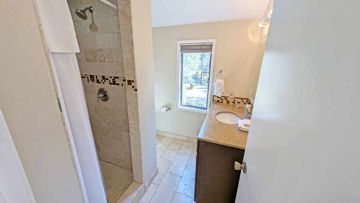 This image features a modern full bathroom with a spacious shower area, a vanity with a sink, and a window view bringing in natural light. The clean lines and calm palette evoke a refreshing, spa-like atmosphere.