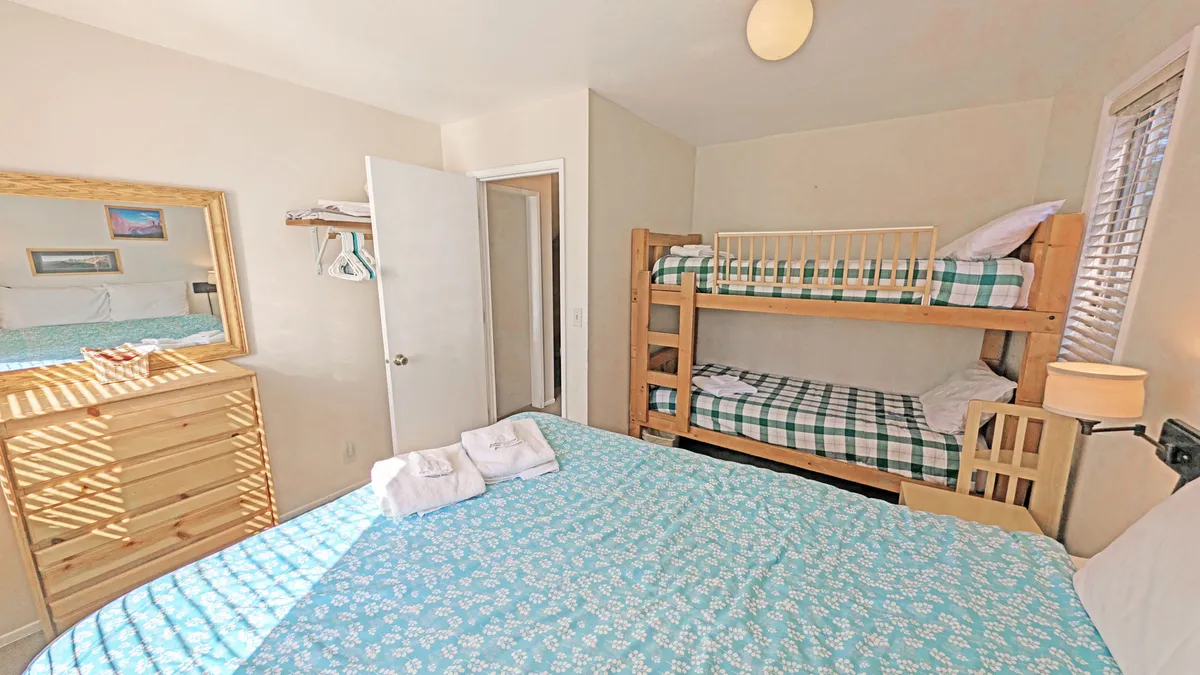 A comfortable bedroom with a bunk bed and queen-sized bed, featuring a dressing mirror, wooden dresser, and colorful bedding. The playful arrangement and inviting decor create a fun yet relaxing atmosphere for children or family groups.